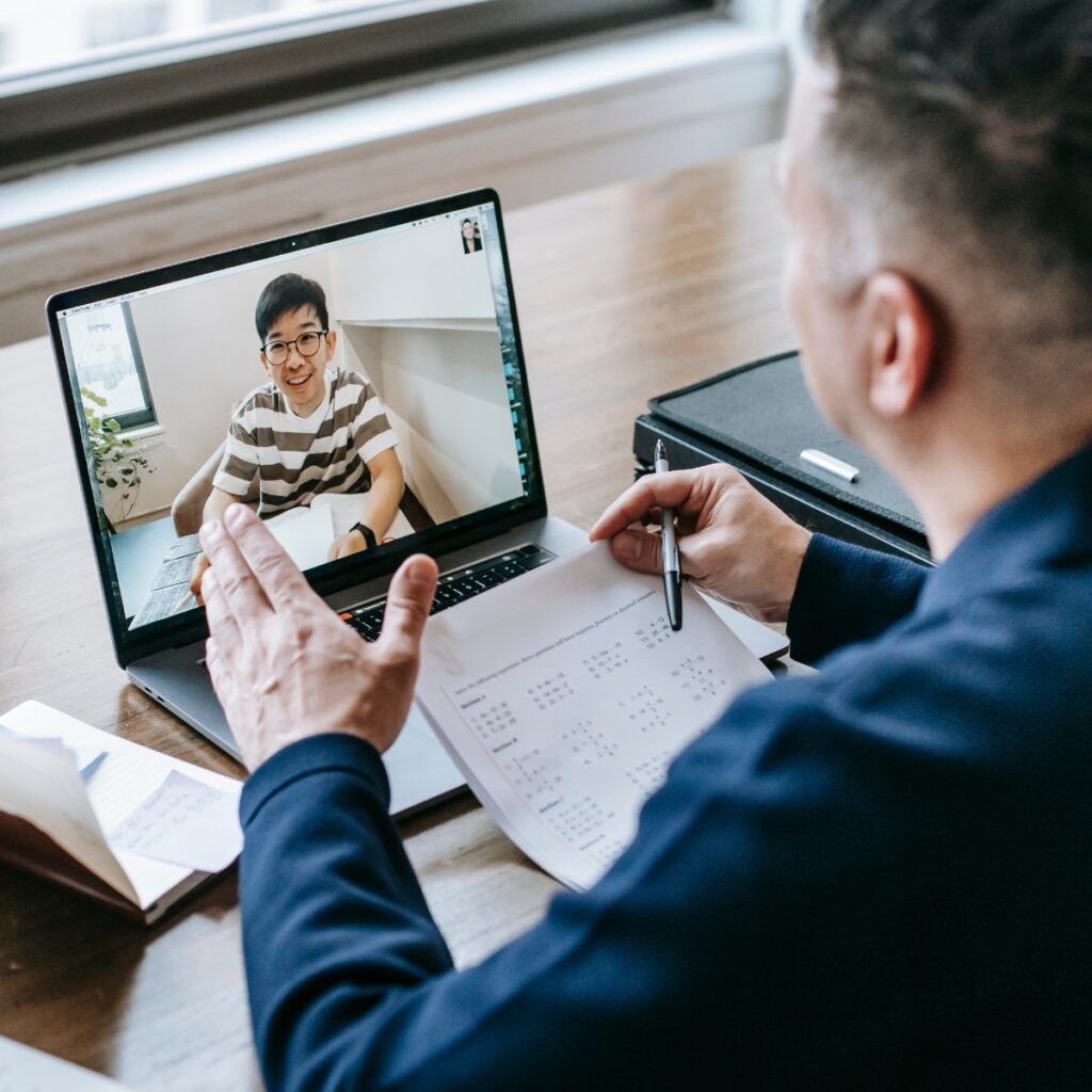 A person takes notes with pen and paper during a video call with their Digital Business Coach. On the laptop screen, the coach wears glasses and a striped shirt, sitting at a desk indoors, ready to discuss online marketing strategies.